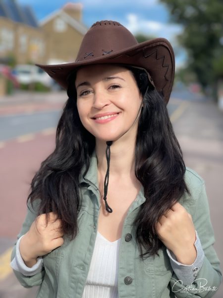 Portrait of a young woman wearing a cowboy hat on a sunny day in London smiling at the camera. The background is blurred by the professional lens. The skin on the face is very refined and the lips are slightly lipsticked to give the photo charm.